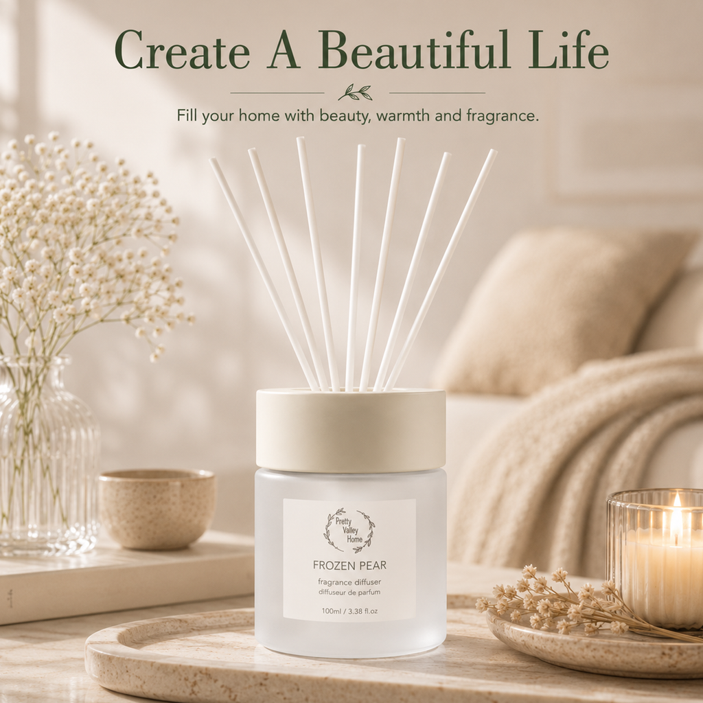 A frosted glass reed diffuser jar labeled 'FROZEN PEAR' sits on a wooden tray in a softly styled home interior. White reeds extend upward from the open top, and a lit candle, dried flowers, and neutral decor surround it.