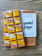 Caramel Frappe soy wax melt snap bar in clear packaging with the Rainy Morning Candles label; caramel-brown wax with yellow and white swirls on a wooden surface.
