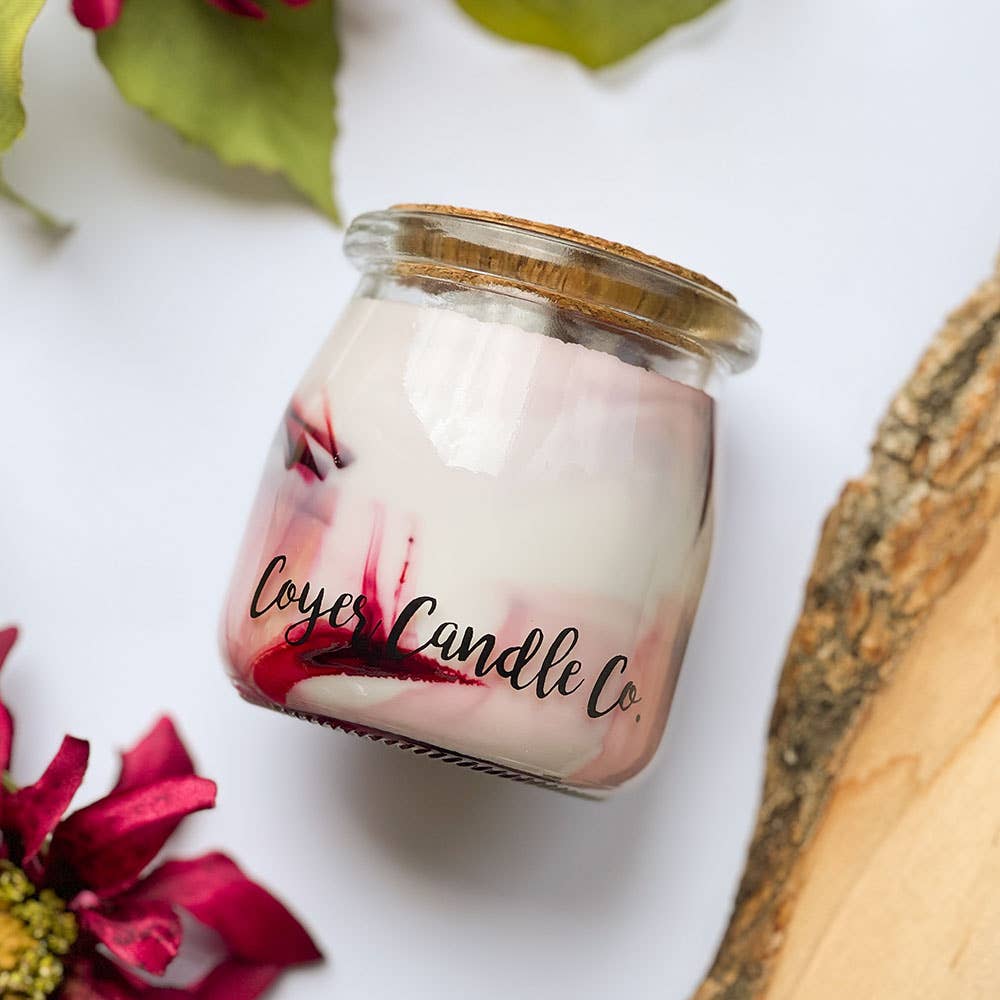 A small glass jar candle with white wax and red marbled swirls, sealed with a cork lid, on a white background with decorative flowers and wood slices. The label reads 'Cozy Candle Co.