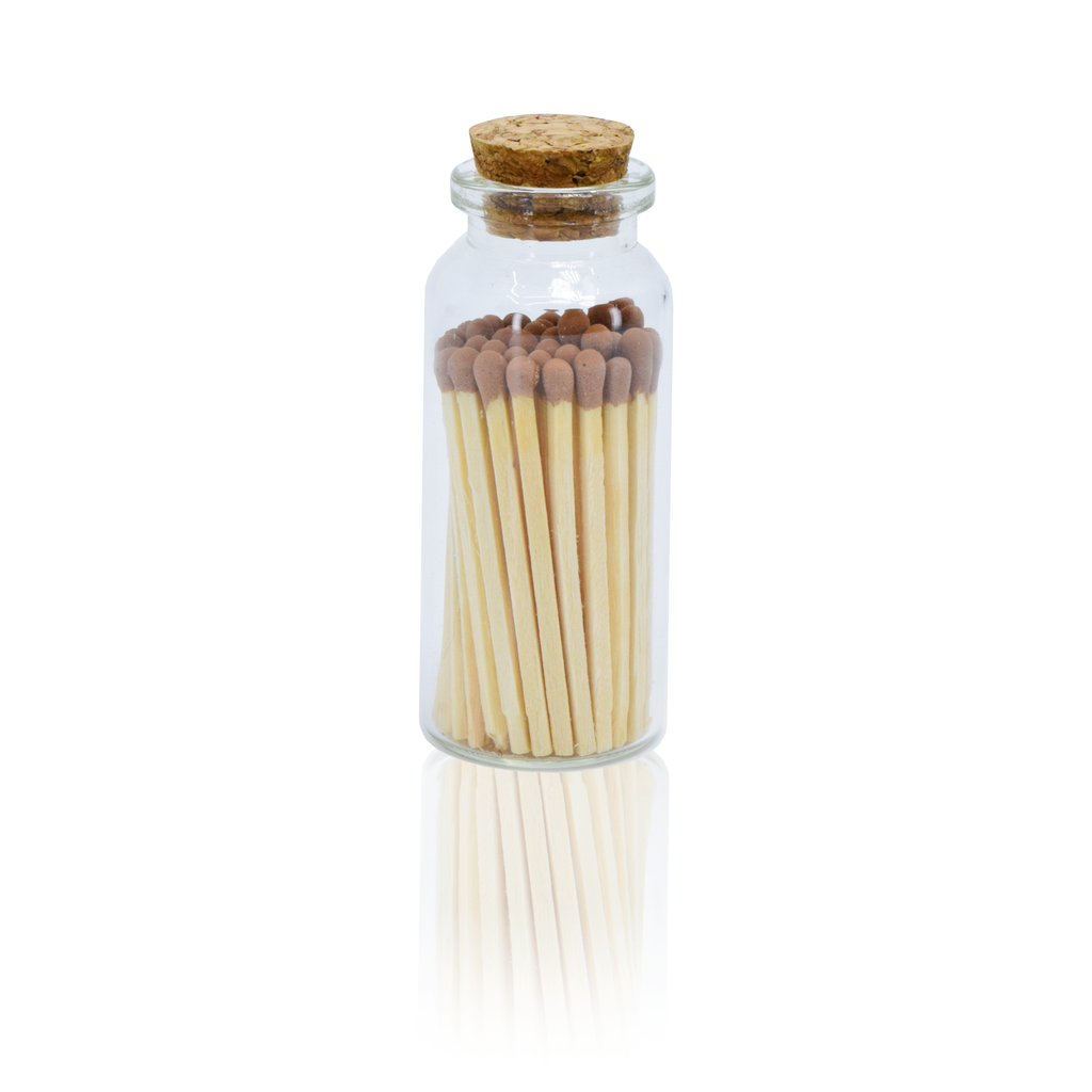 A clear glass bottle with a cork stopper containing a set of wooden matches with brown tips, shown on a white background with a soft reflection below.
