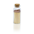 A clear glass bottle with a cork stopper containing a set of wooden matches with brown tips, shown on a white background with a soft reflection below.