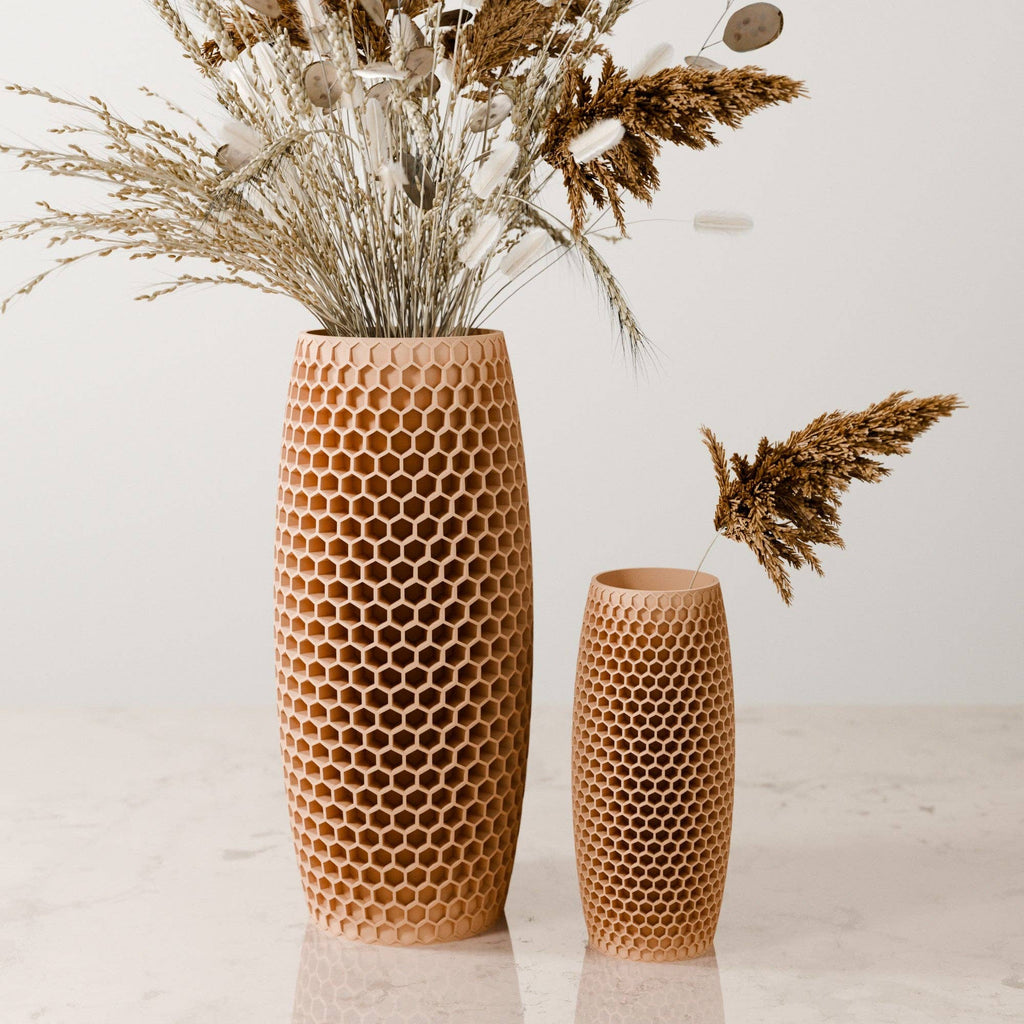 Two honeycomb-patterned wooden vases in different heights, filled with dried grasses and botanicals, set on a light marble surface against a plain off-white background.