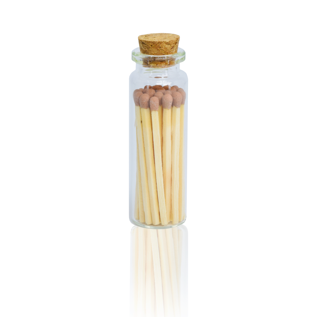 A small clear glass bottle with a cork stopper containing a set of wooden matches with brown tips, centered on a white background with a faint reflection below.