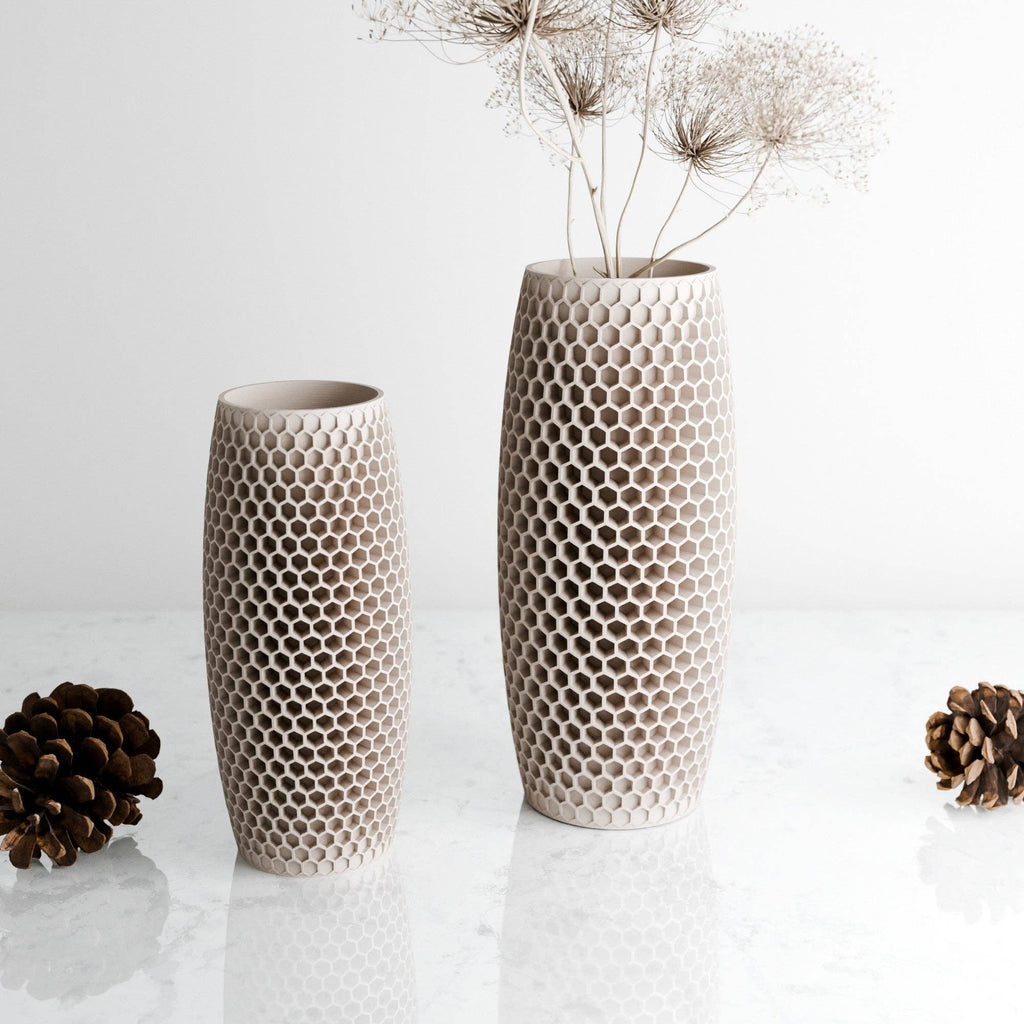 Two textured plastic vases with a honeycomb pattern, one short and one tall, displayed on a white marble surface with pinecones and dried grass stems.