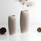 Two textured plastic vases with a honeycomb pattern, one short and one tall, displayed on a white marble surface with pinecones and dried grass stems.