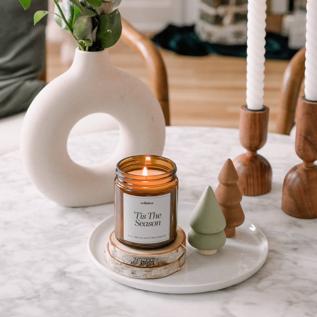 Candle labeled 'Tis The Season' on a marble surface with decorative items and a vase in the background.