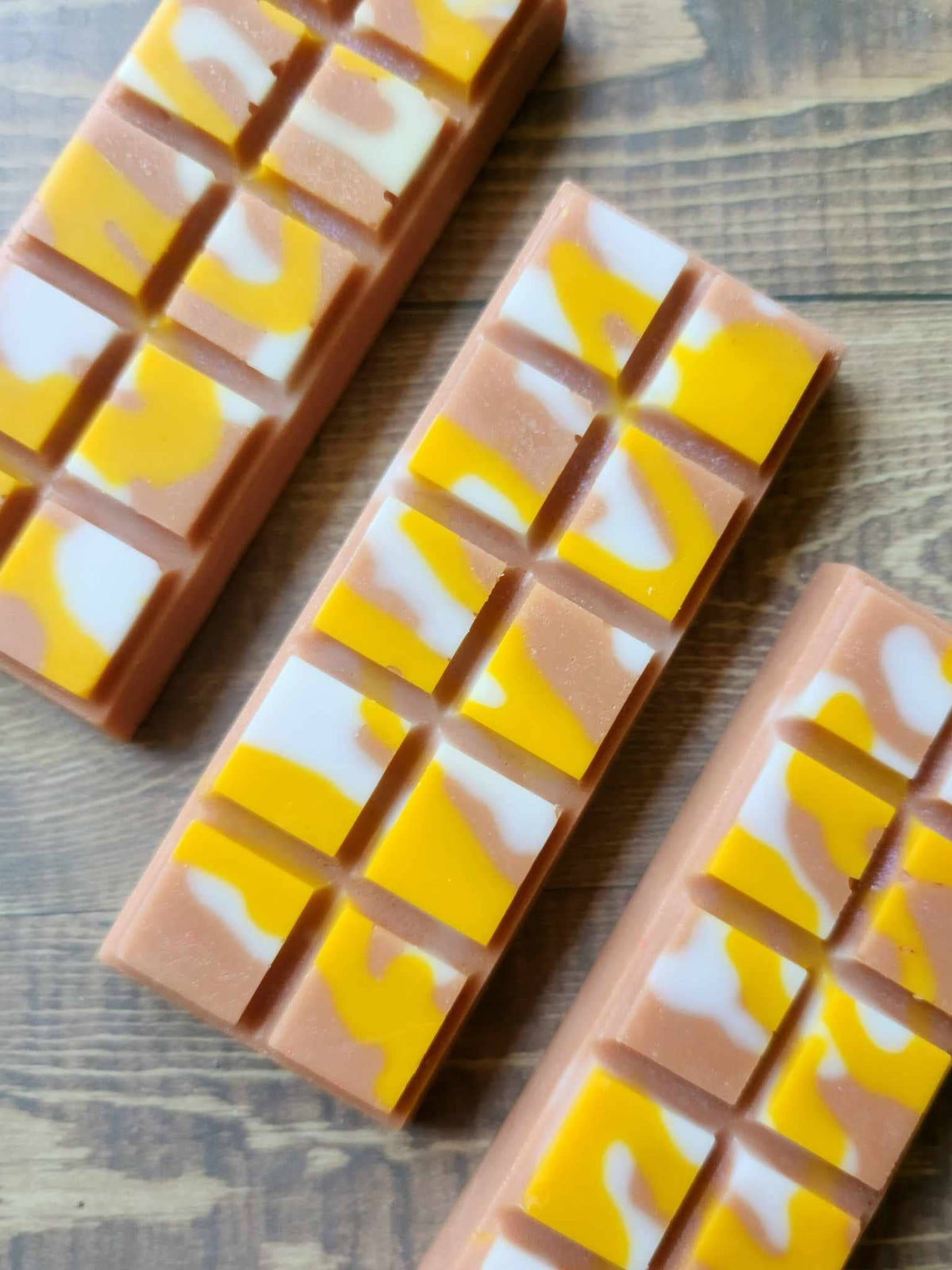 Three unwrapped Caramel Frappe soy wax melt snap bars, caramel-brown wax with yellow and white swirls, on a wooden surface.