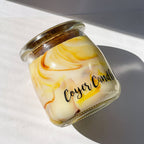 A small glass jar candle with a marbled cream and yellow wax design and a gold-toned metal lid, photographed on a white surface with strong sunlight and shadows. The label reads 'Cozy Candle'.