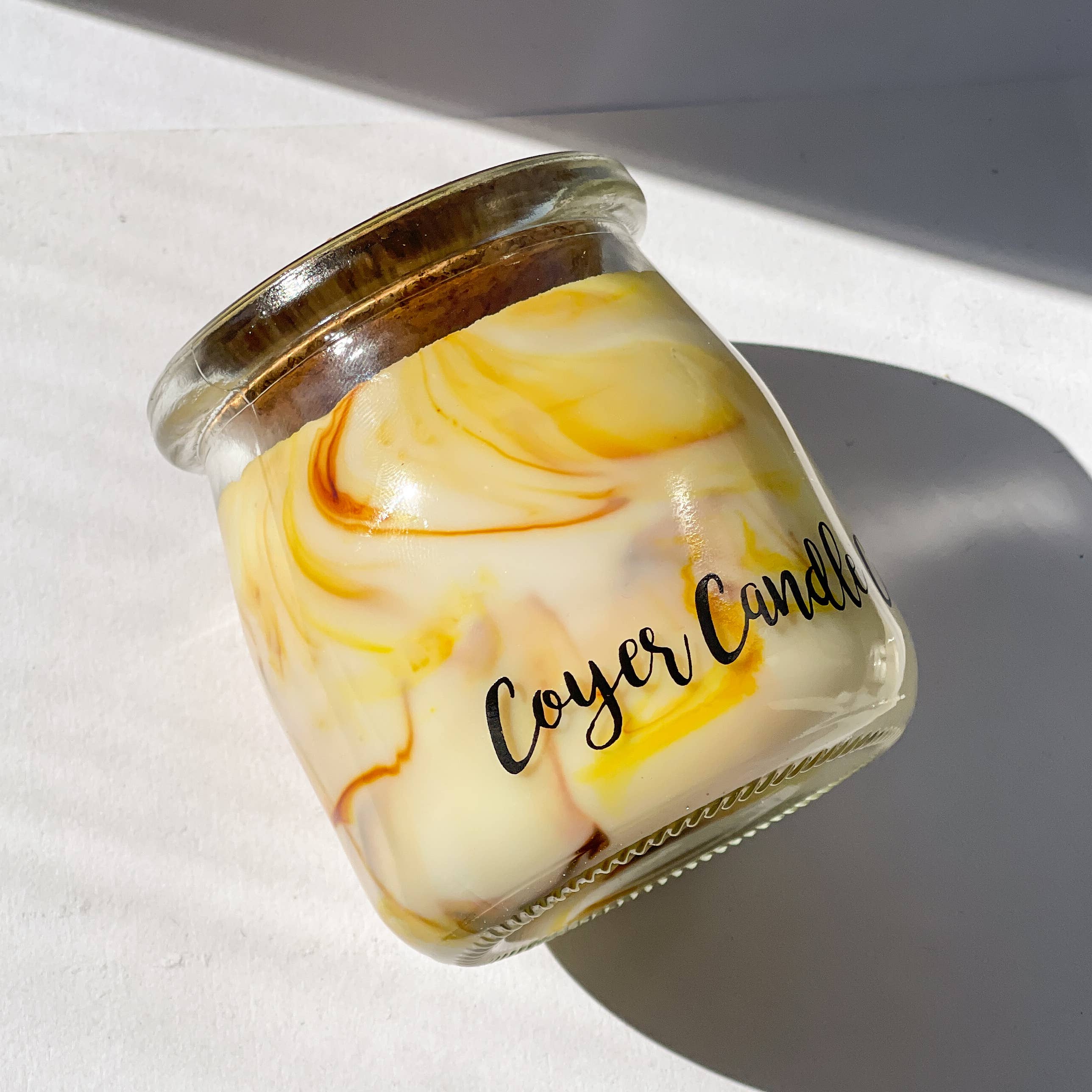 A small glass jar candle with a marbled cream and yellow wax design and a gold-toned metal lid, photographed on a white surface with strong sunlight and shadows. The label reads 'Cozy Candle'.