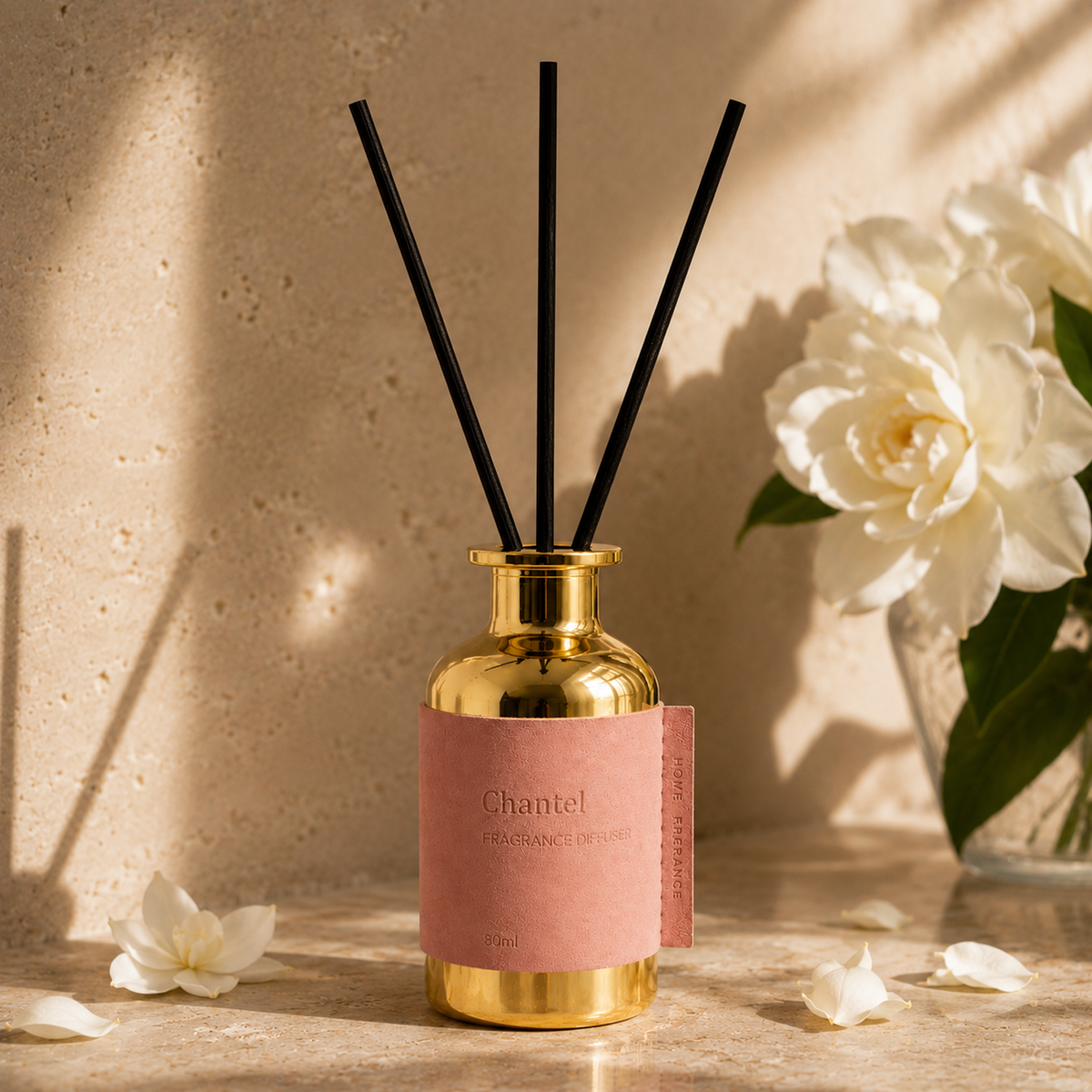 Gold diffuser bottle with pink label and black reeds on a wooden surface with white flowers.