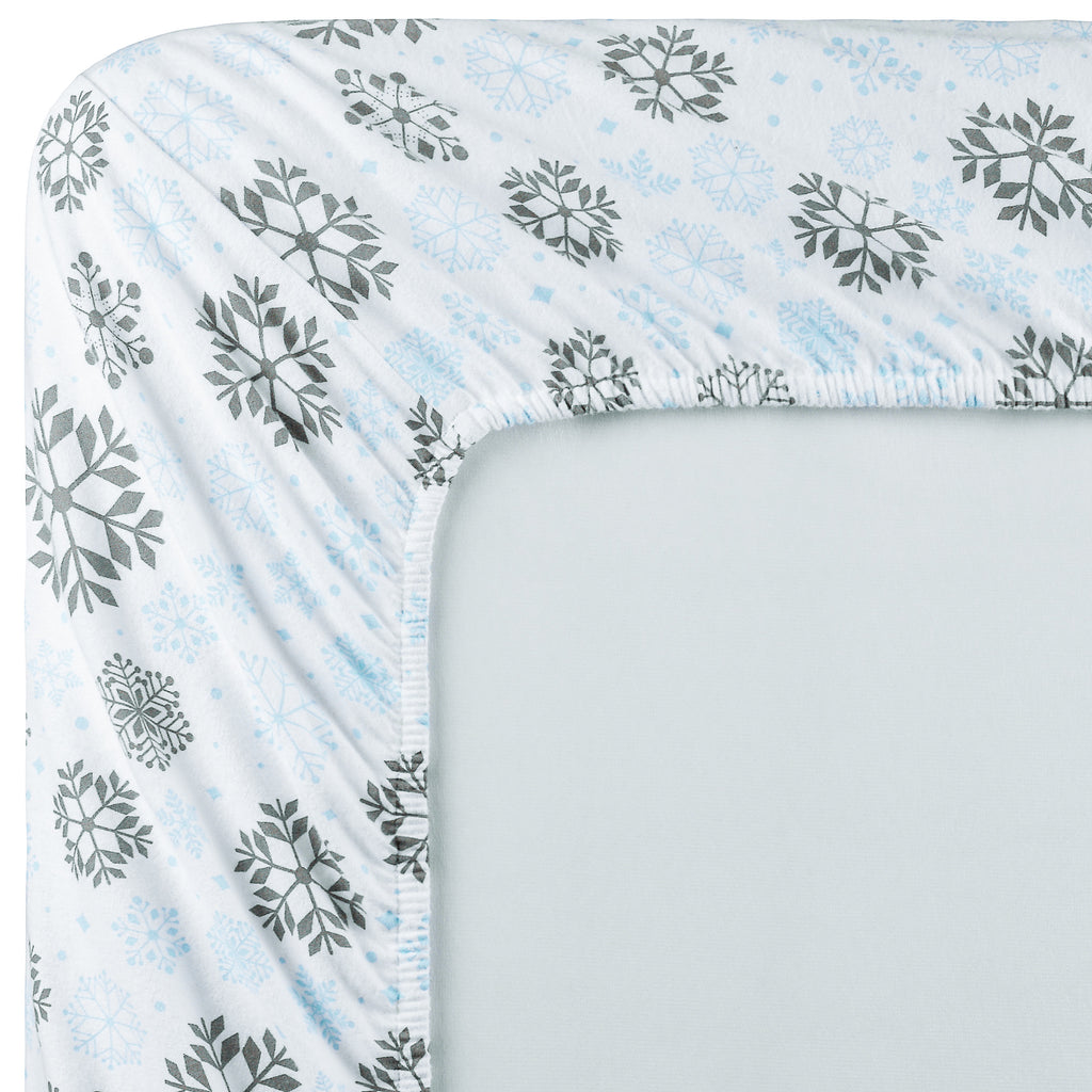A close-up of a fitted sheet corner showing a white base with a pattern of gray and light blue snowflakes, fitted around the edge of a mattress.