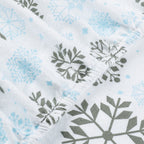 Close-up of a white fabric item, likely a baby blanket or swaddle, featuring a repeating pattern of light blue snowflakes and dark gray stylized leaf or pine branch motifs.