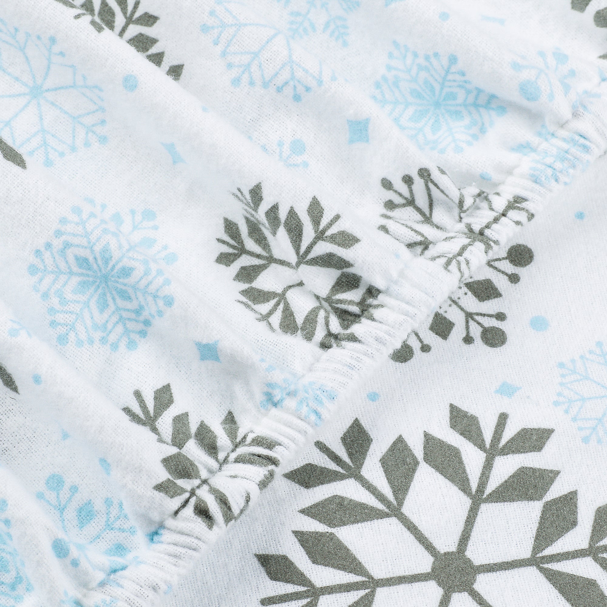 Close-up of a white fabric item, likely a baby blanket or swaddle, featuring a repeating pattern of light blue snowflakes and dark gray stylized leaf or pine branch motifs.