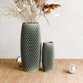 Two textured sage green ceramic vases with a honeycomb pattern, one tall and one short, styled with dried grasses on a light wood surface against a neutral wall.