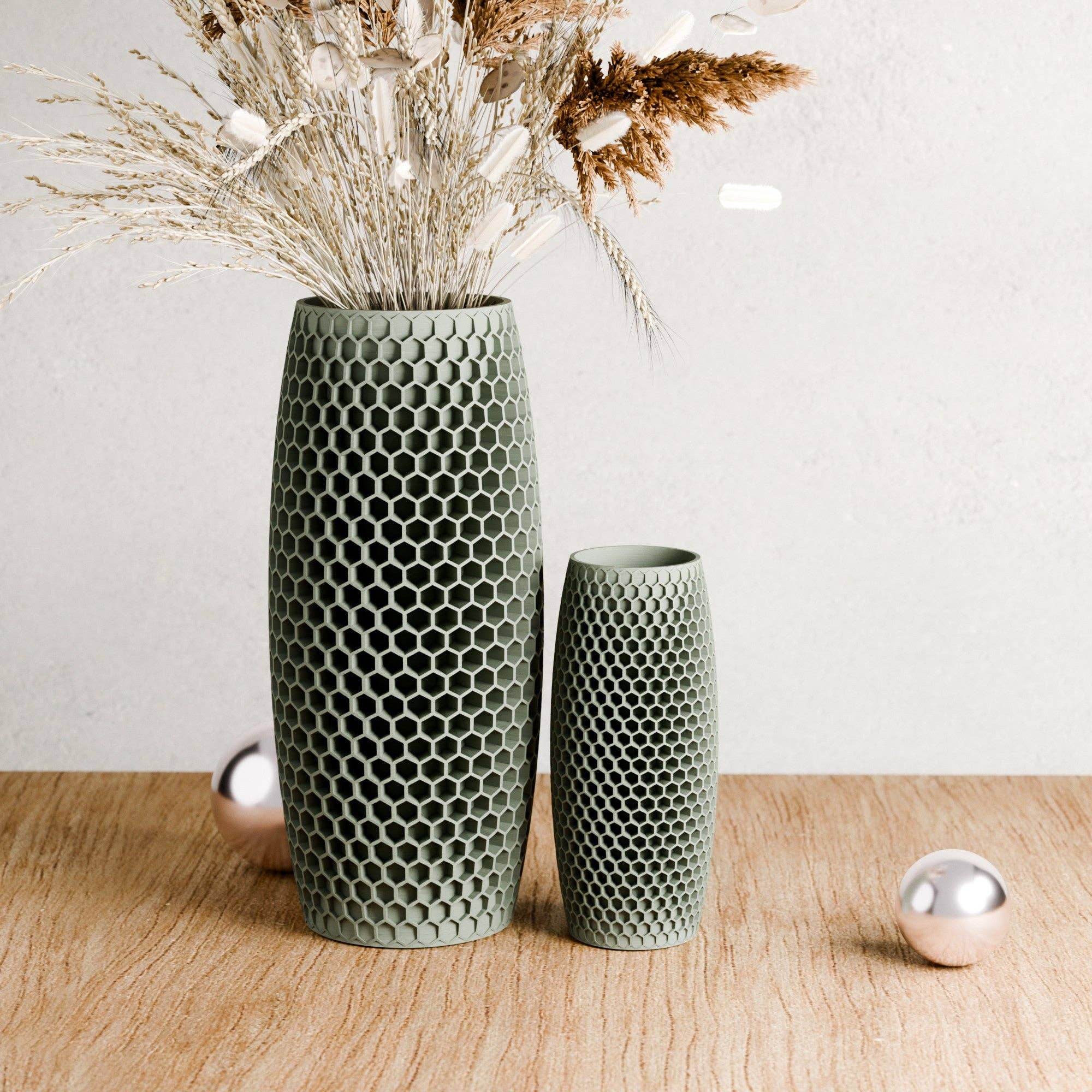 Two textured sage green ceramic vases with a honeycomb pattern, one tall and one short, styled with dried grasses on a light wood surface against a neutral wall.