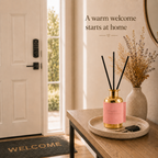 A pink reed diffuser bottle with gold accents and black reeds sits on a stone tray on a wooden console table in a warmly lit entryway. A beige vase with dried flowers, a white door, and the text 'A warm welcome starts at home' are visible.