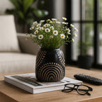 A black ceramic vase with beige geometric patterns, filled with small white daisy flowers, styled on a wooden table next to a remote control and glasses.