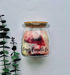 A glass jar candle with a cork lid and a visible 'Loyer Candle Co.' label, filled with cream wax and pink-red marbled swirls, photographed against a light gray background with greenery on the left.