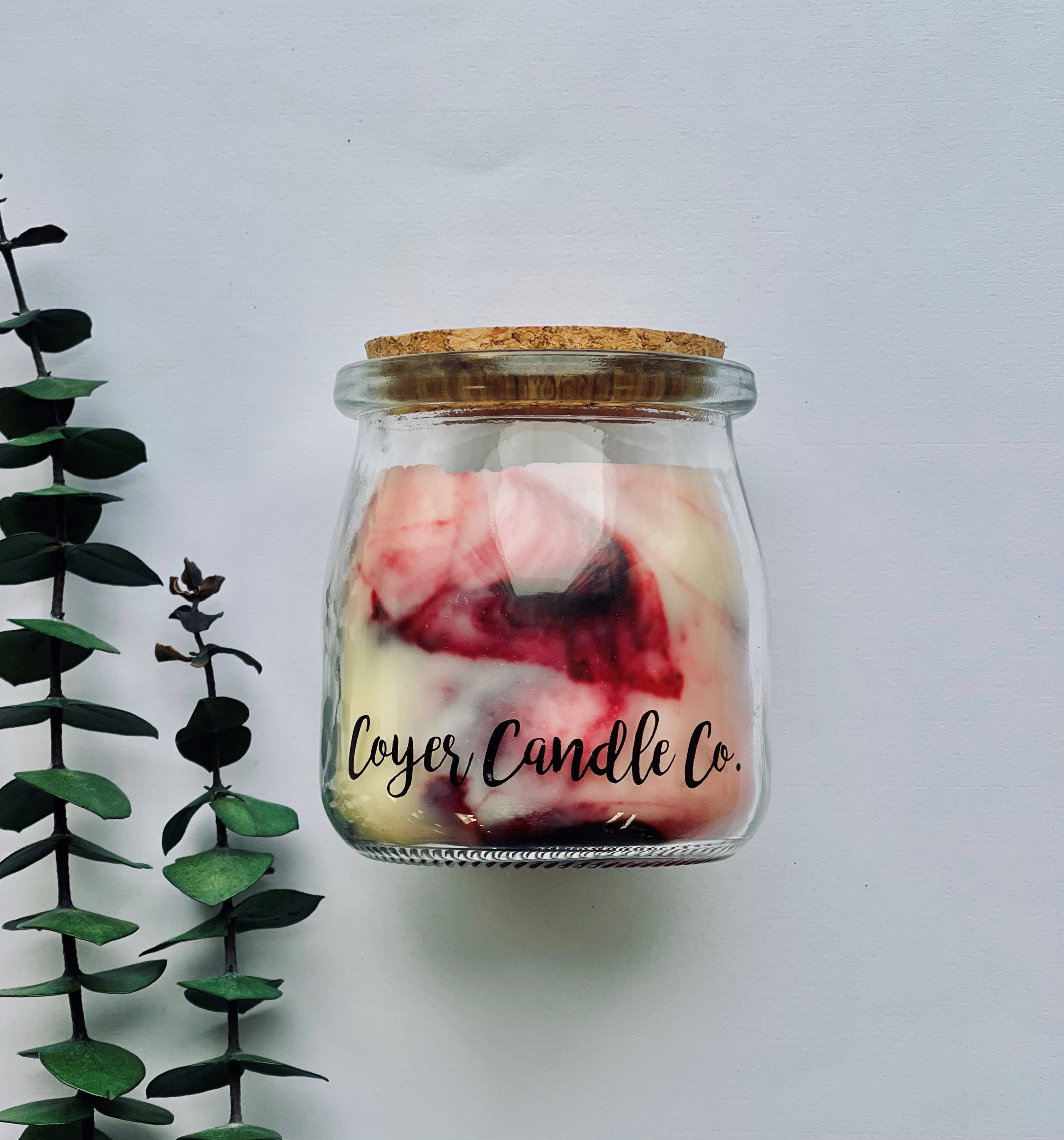 A glass jar candle with a cork lid and a visible 'Loyer Candle Co.' label, filled with cream wax and pink-red marbled swirls, photographed against a light gray background with greenery on the left.