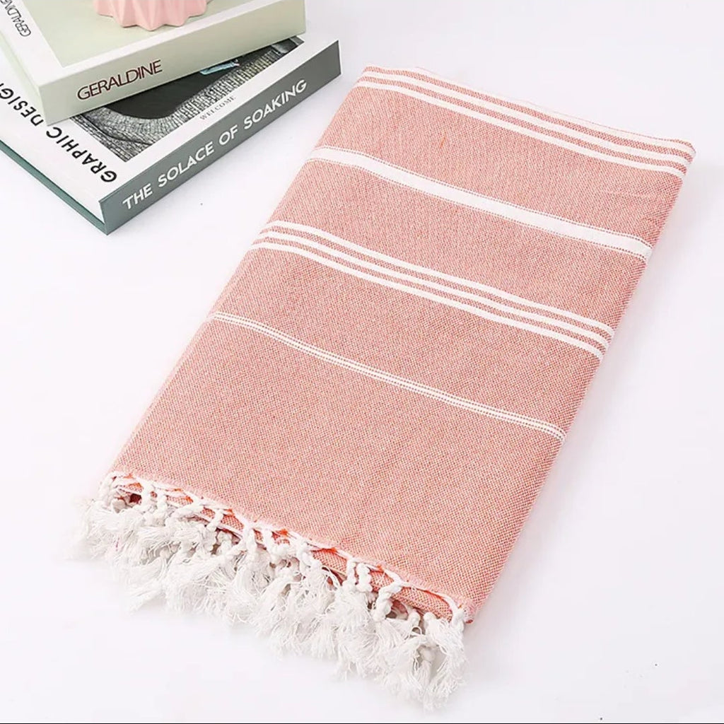 Folded coral pink Turkish beach towel with white stripes and tassels beside a stack of books