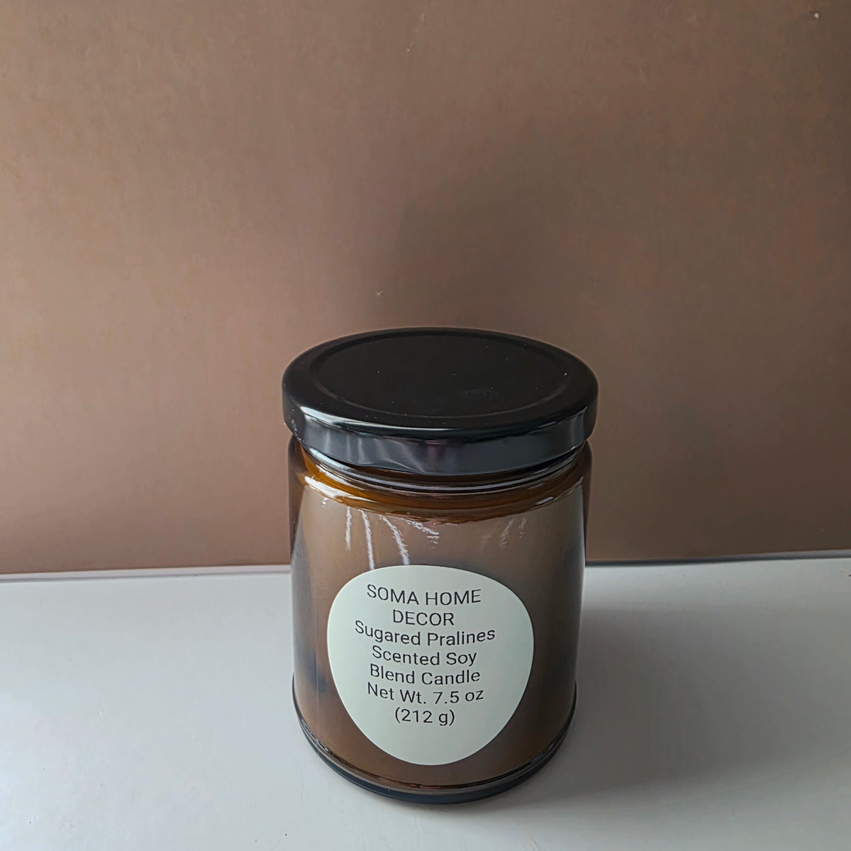 A 7.5 oz scented soy candle in an amber glass jar with a black lid and a white circular label reading 'SOMA HOME DECOR Sugared Pralines Scented Soy Blend Candle'.