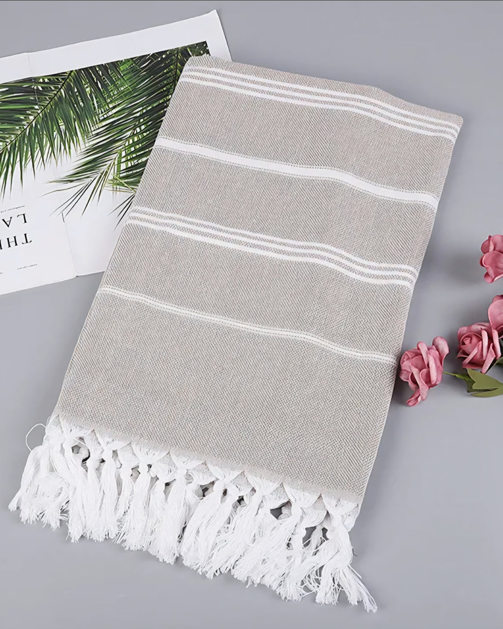 Tan Turkish beach towel with white stripes and knotted tassels, styled with a magazine and pink roses