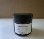 A handpoured soy candle in a dark amber glass jar with a black lid and a white label, set against a plain brown background on a light surface.