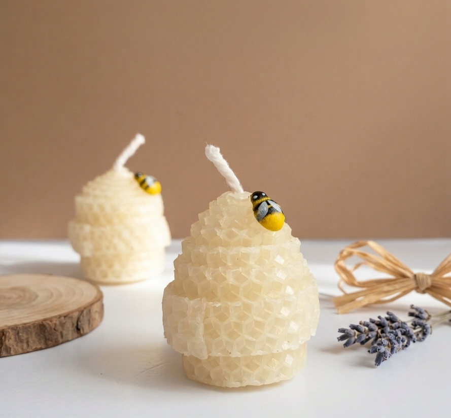 Two honeycomb-style beeswax candles with white wicks and small bumblebee decorations, styled on a white surface with a wood slice, lavender sprigs, and a dried bow against a warm beige background.