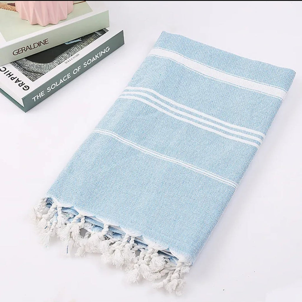 Folded light blue Turkish beach towel with white stripes and tassels beside a stack of books