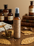 A bottle of Tobacco Linen & Room Spray stands on a wooden table surrounded by dried tobacco leaves, a brass mortar and pestle, and vintage-style amber apothecary bottles with labels such as Cedar, Tang-Yang, and Amber.