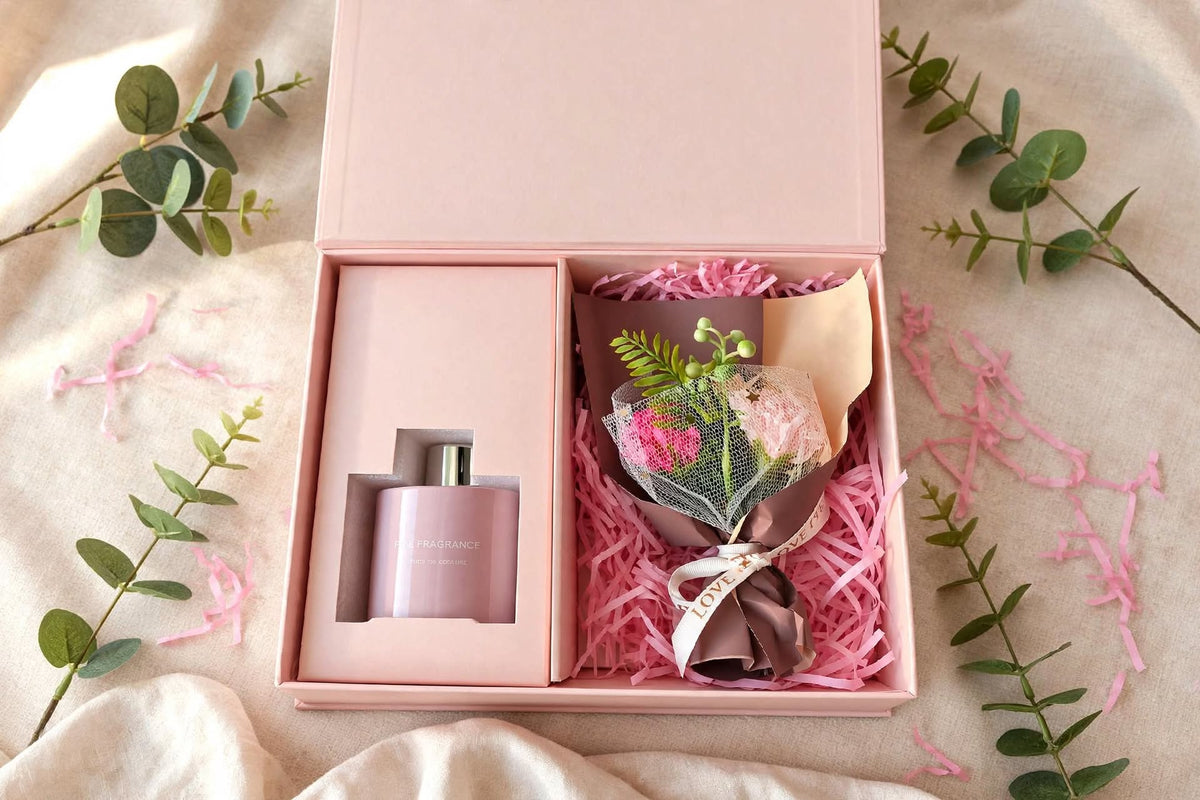 Reed diffuser gift set with bouquet: open pink gift box with home fragrance diffuser and pink flower bouquet on pink shredded paper, styled with eucalyptus