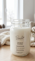 A handpoured soy candle in a clear glass mason jar sits on a light wood surface near a window. The label reads 'Soma Home' and 'Soft Wool & Snowflakes.