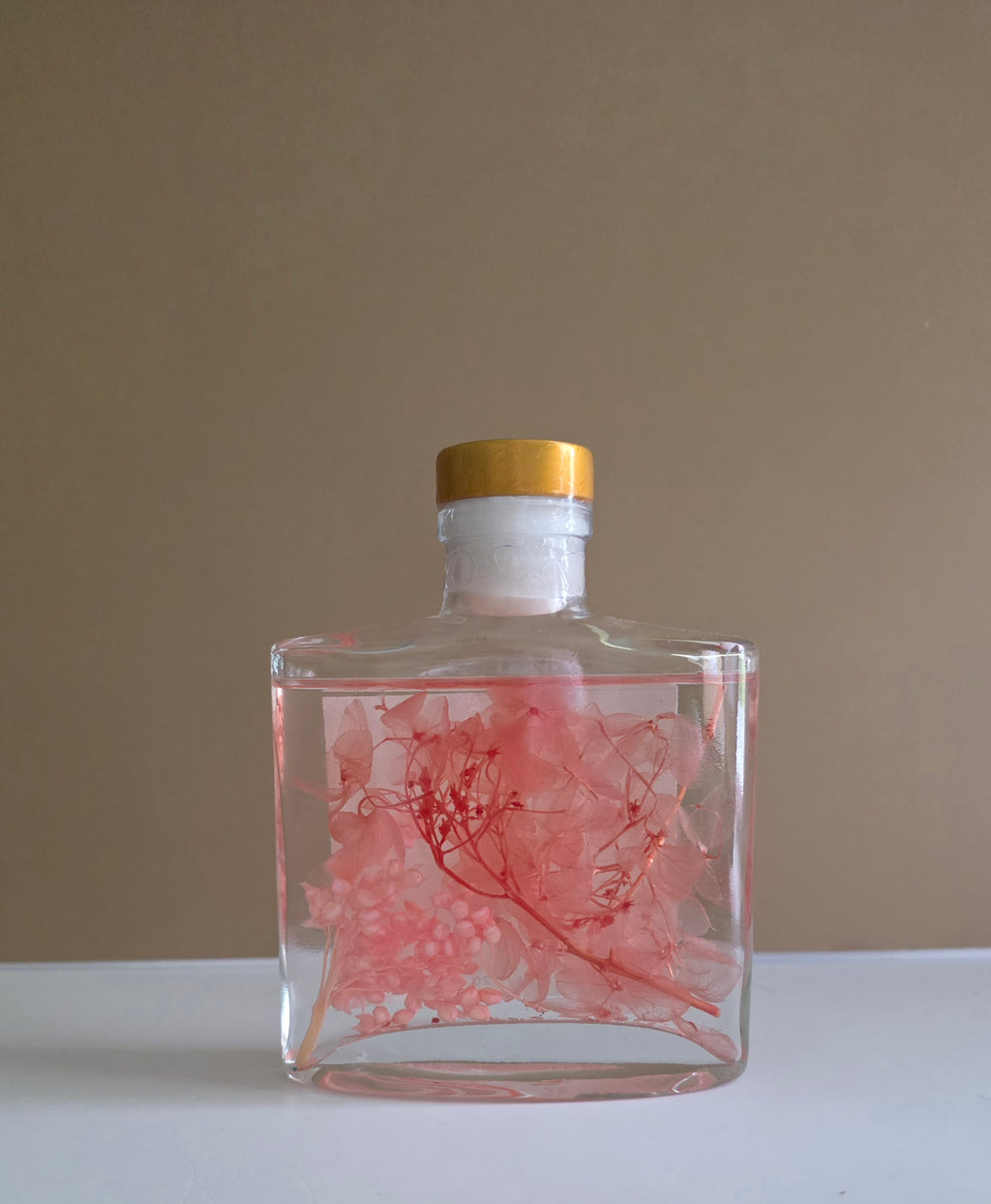 A clear glass bottle with a gold cap, filled with pink liquid and delicate dried floral elements, set against a plain beige background.