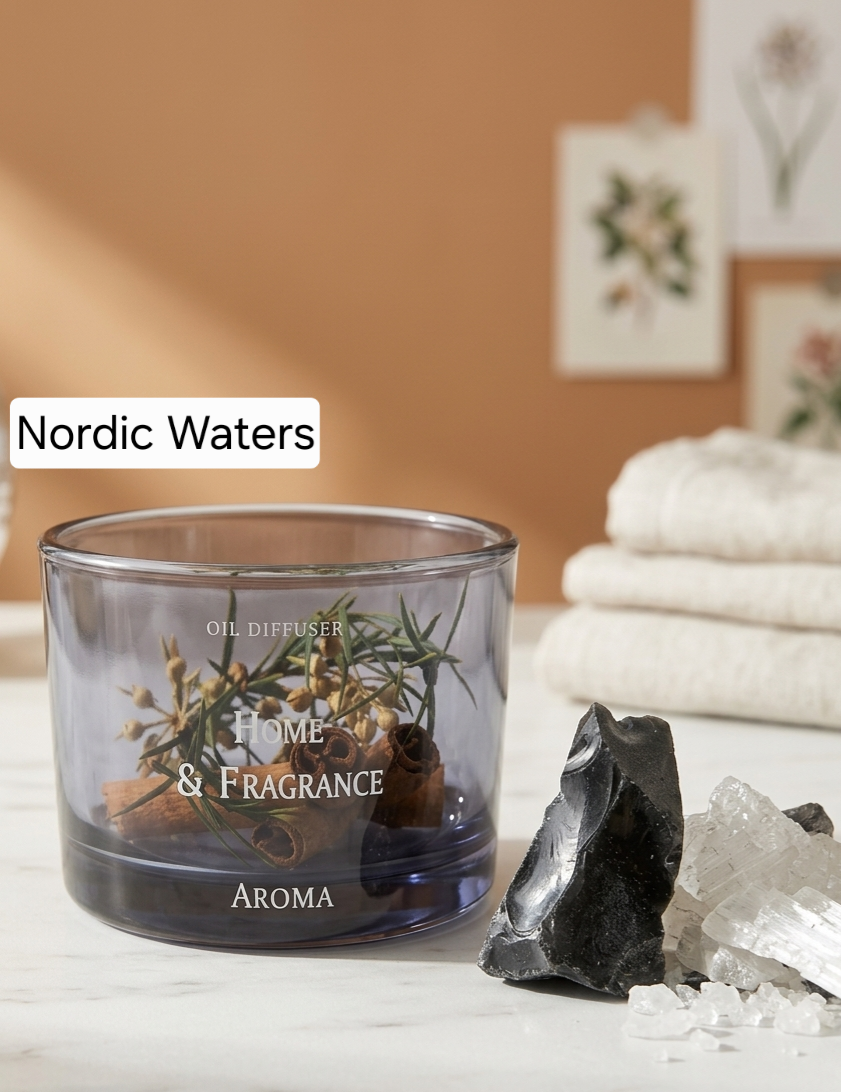 A glass oil diffuser candle labeled 'Nordic Waters' with 'Home & Fragrance' and 'AROMA' printed on the front, filled with botanicals and rolled paper. The diffuser sits on a marble surface with crystals and folded towels in the background against a warm terracotta wall.