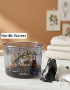 A glass oil diffuser candle labeled 'Nordic Waters' with 'Home & Fragrance' and 'AROMA' printed on the front, filled with botanicals and rolled paper. The diffuser sits on a marble surface with crystals and folded towels in the background against a warm terracotta wall.