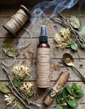 A bottle of Aldo Santo Linen & Room Spray stands on a rustic wooden surface surrounded by natural props including dried flowers, twine, branches, a spoon, and a rolled paper scroll. Smoke curls around the bottle, and the label is clearly visible.
