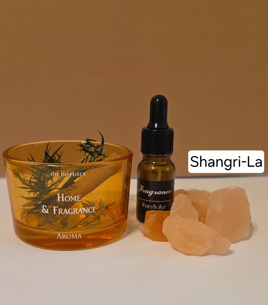 A home fragrance set featuring an amber glass oil diffuser candle with rosemary sprigs, a small amber dropper bottle labeled 'Fragrance Fresh Air', and pink salt crystals on a white surface against a warm brown background. A text label reads 'Shangri-La'.