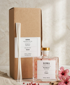 SOMA Home Decor Botanical Scent Coco reed diffuser set, featuring a clear glass bottle with a gold cap, white reeds, a brown kraft cardboard box, and pink flowers on a neutral textured background.