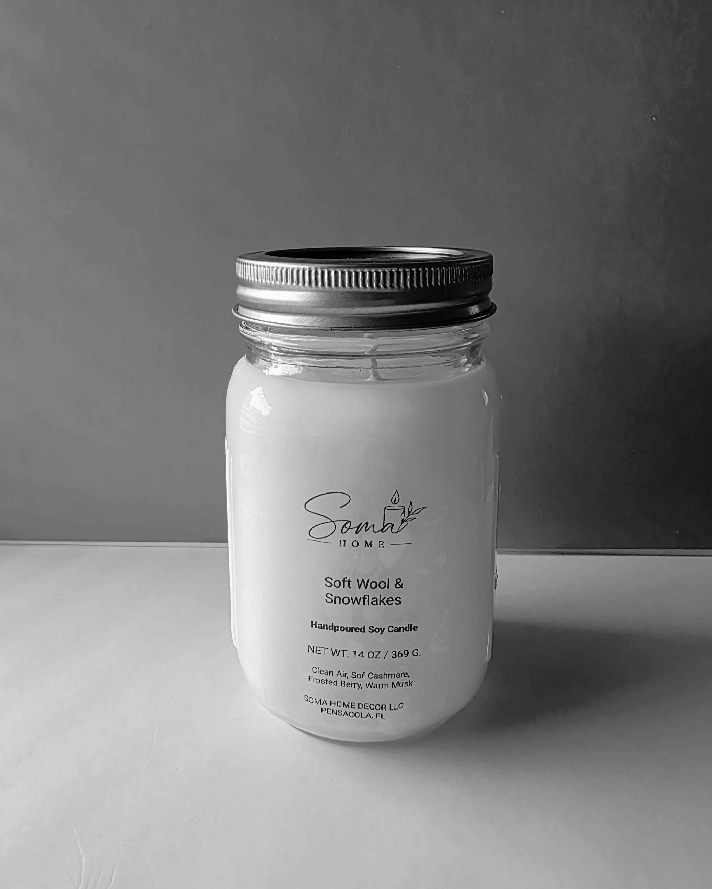 A handcrafted soy candle in a clear glass mason jar with a silver metal lid, featuring a white wax fill and a printed label that reads 'Scent of Home' and 'Soft Wool & Snowflakes'. The candle is photographed on a light gray surface against a darker gray background.
