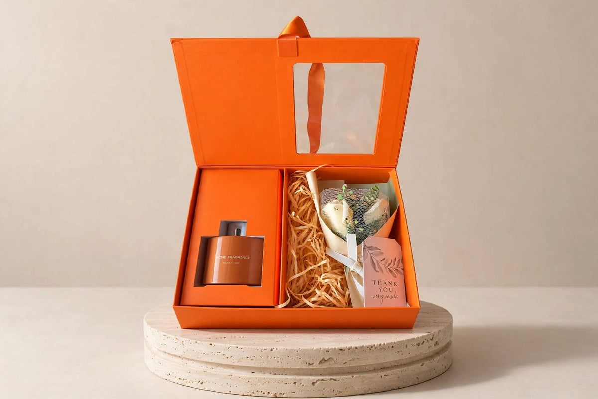Reed diffuser gift set with bouquet: open orange gift box with diffuser and white rose bouquet on a stone pedestal