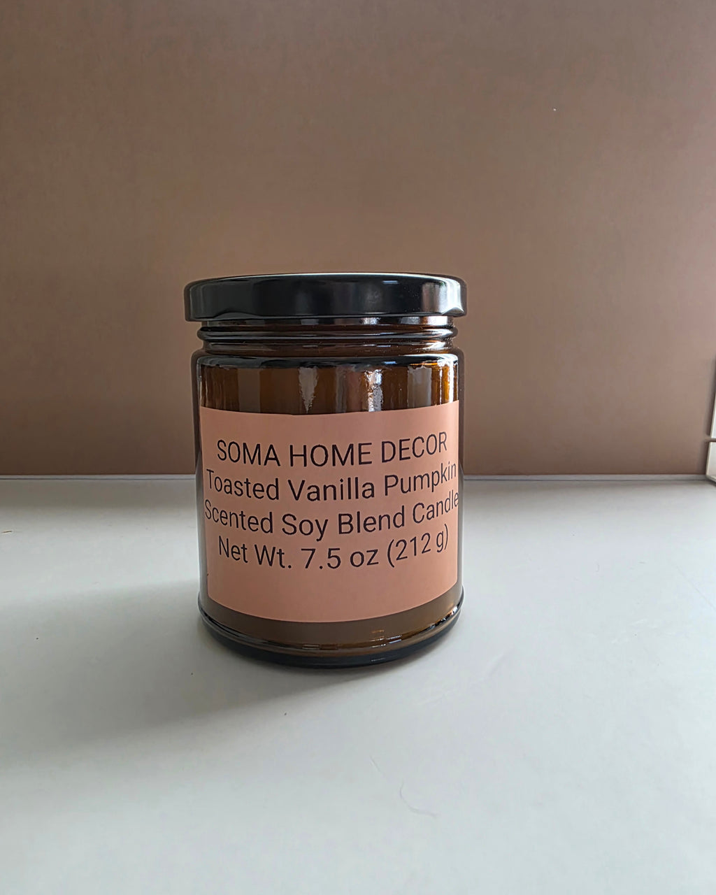A 7.5 oz amber glass jar candle with a black lid and a peach-colored label reading 'SOMA HOME DECOR Toasted Vanilla Pumpkin Scented Soy Blend Candle'.