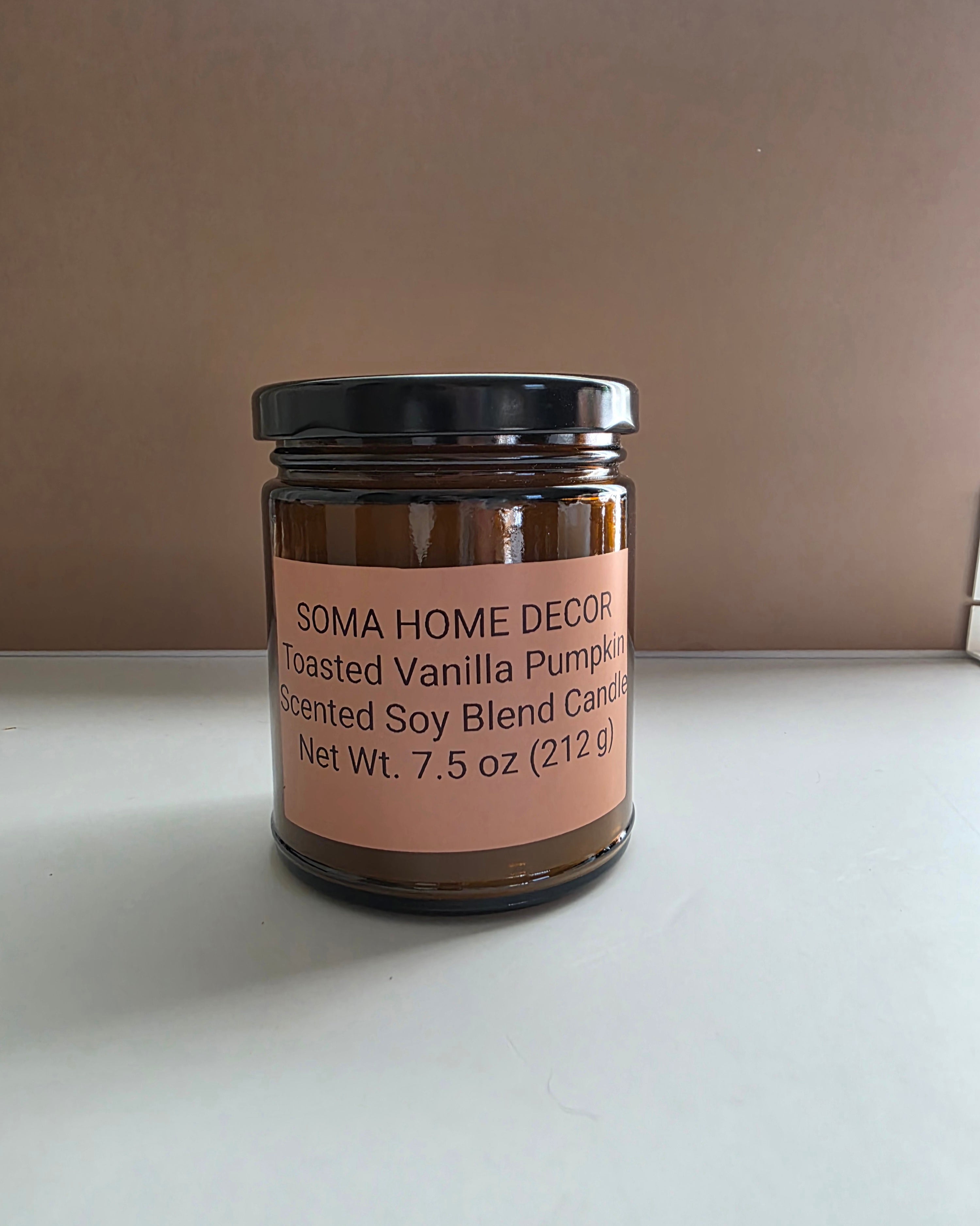 A 7.5 oz amber glass jar candle with a black lid and a peach-colored label reading 'SOMA HOME DECOR Toasted Vanilla Pumpkin Scented Soy Blend Candle'.