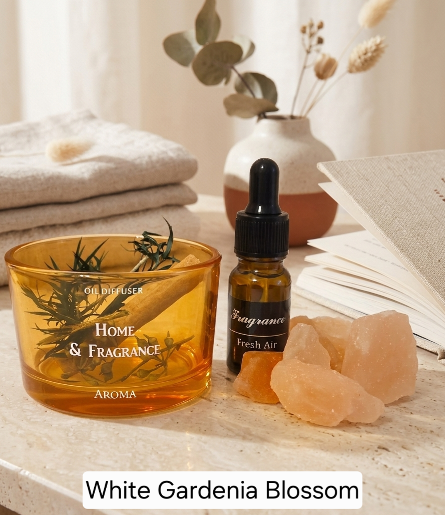 A home fragrance product set on a light stone surface, featuring an amber glass oil diffuser bowl labeled 'HOME & FRAGRANCE' and 'AROMA', a small amber dropper bottle labeled 'Fragrance Fresh Air', pink salt crystals, folded beige towels, a vase with dried stems, and an open book. Text at the bottom reads 'White Gardenia Blossom'.