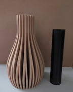 A large sculptural vase made of vertically slatted wood in a light natural tan color, shown next to a simple matte black cylindrical vase on a white surface against a warm brown wall.