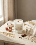 A Soma Home Decor Cashmere Cedar scented soy blend candle in a clear glass jar sits on a light stone table styled with a cream throw blanket, dried wheat stalks, wooden cubes, and star anise in a warm, natural setting.