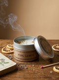 A pink lemon scented candle in a silver metal tin with its lid off, showing rising smoke. The tin is labeled 'PINK LEMON' and sits on a rustic wooden table surrounded by dried orange slices, lavender buds, a match, and a book.