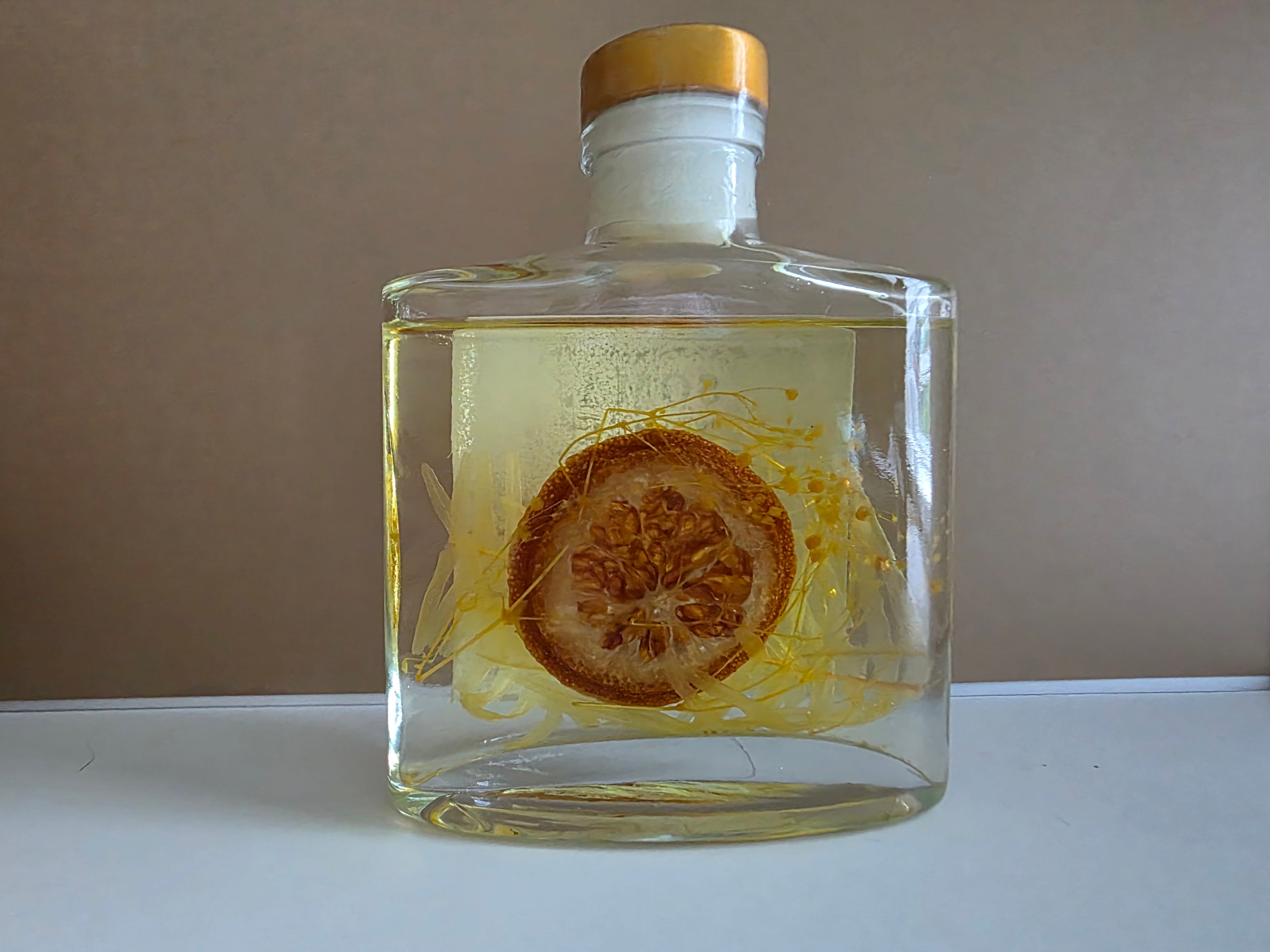 A clear glass bottle with a gold cap, filled with a pale yellow liquid and containing a dried citrus slice and fine golden threads, set against a plain beige background.