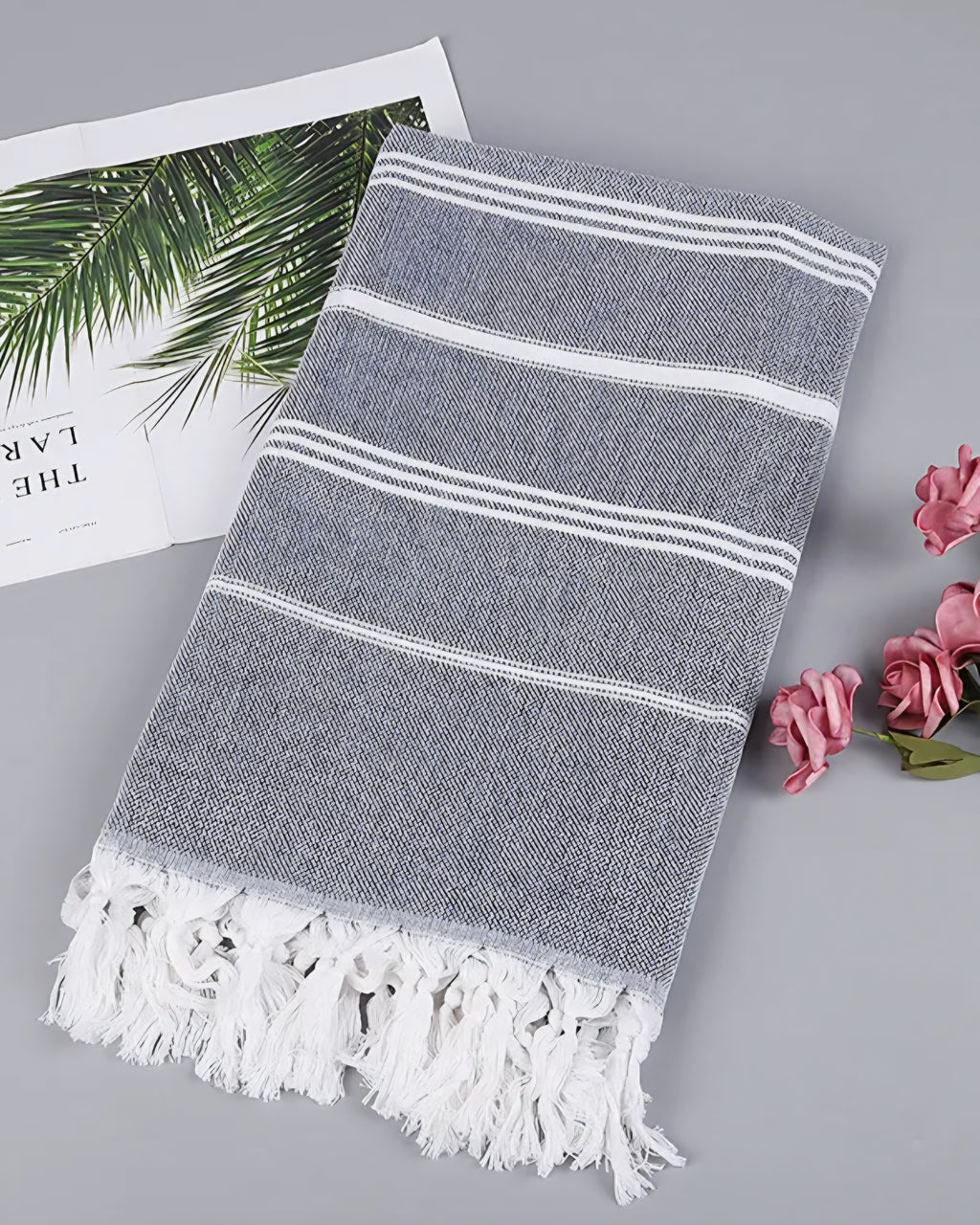 Dark gray Turkish beach towel with white stripes and knotted tassels, styled with a magazine and pink roses
