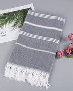 Dark gray Turkish beach towel with white stripes and knotted tassels, styled with a magazine and pink roses