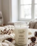 A handpoured soy candle in a clear glass jar sits on a light wood surface wrapped in a cream knit blanket, with pinecones and dried botanicals around it. The candle label reads 'Soma Home' and 'Soft Wool & Snowflakes'.